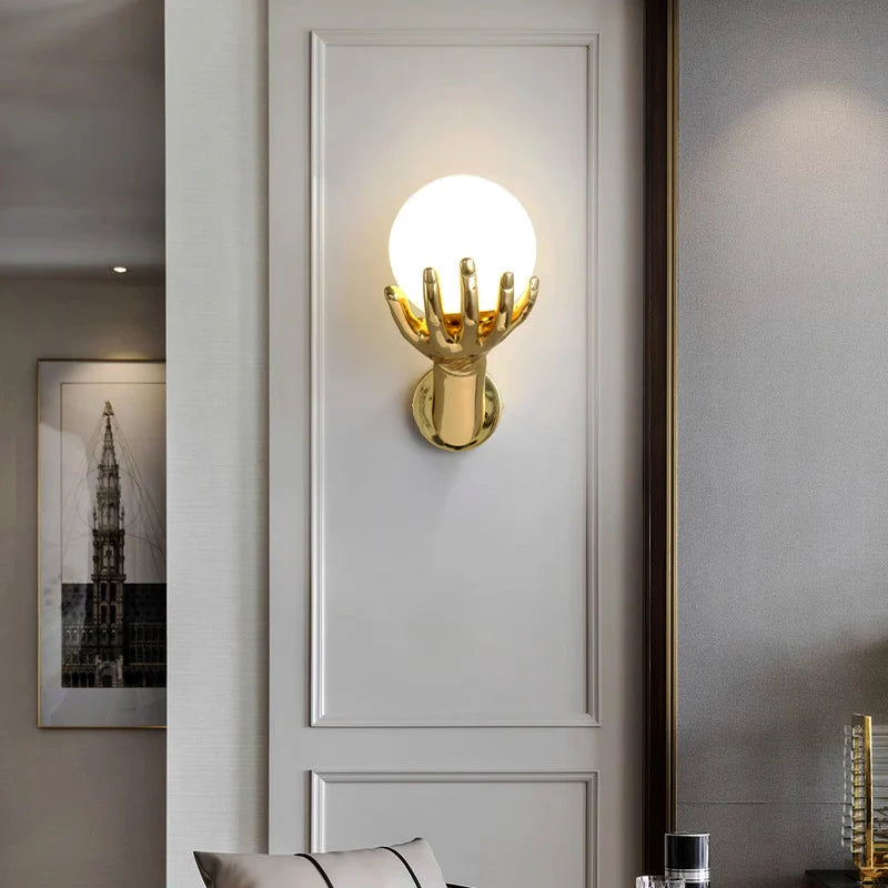 The golden hand-shaped wall lamp illuminates the white wall area, with decorative paintings and ornaments next to it.