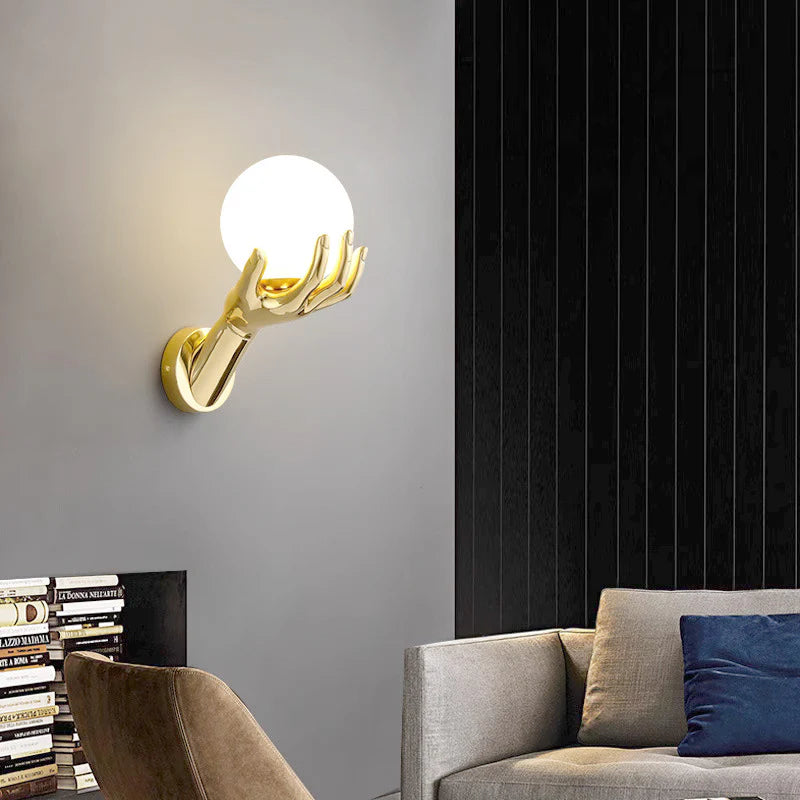 The golden hand-shaped wall lamp lights up the gray wall, and the soft light falls down. There are sofas, pillows and stacked books on the side.