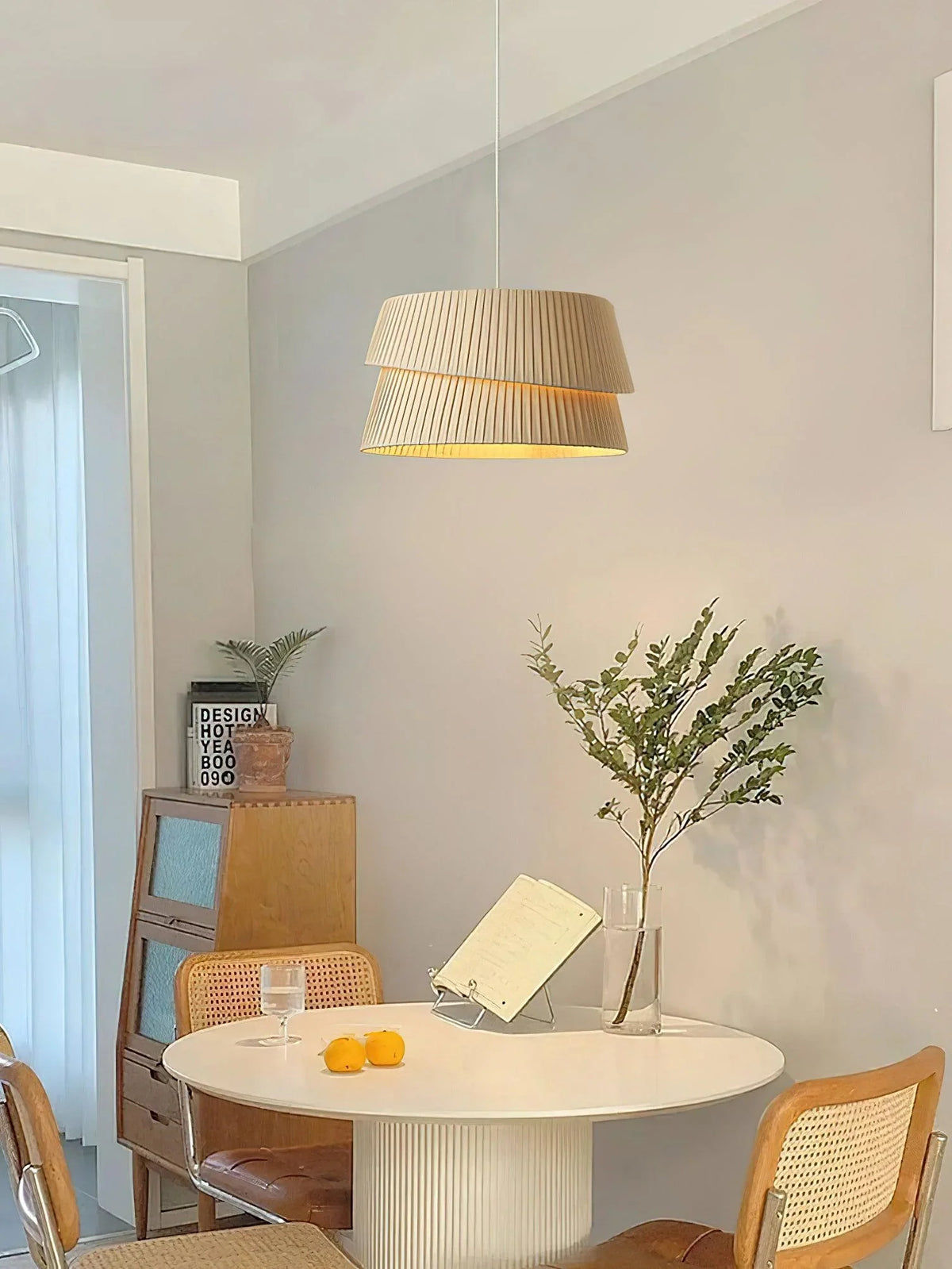 Indoors, in front of the light grey wall, a beige double-layer pleated-textured chandelier hangs above the white round table, providing soft lighting.
