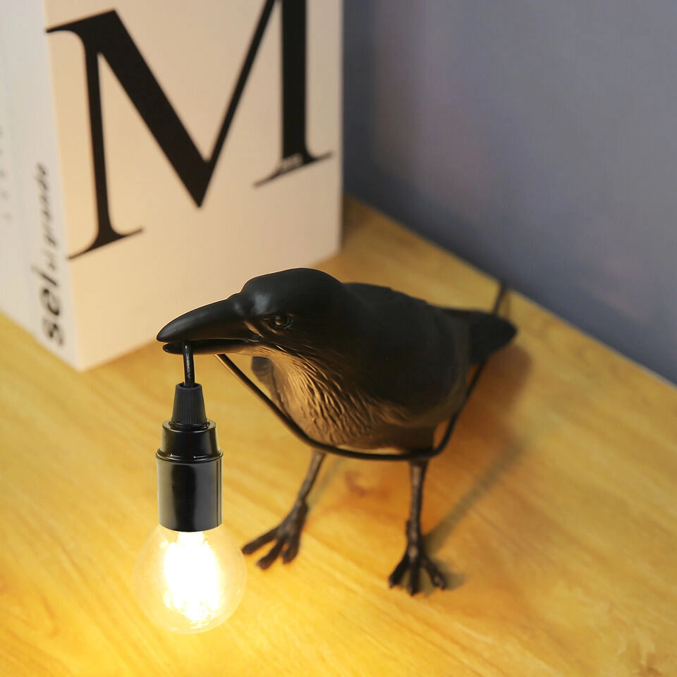 A black crow-shaped lamp, the crow's beak holds the connecting wire of the light bulb, and the light bulb is lit with warm yellow light.