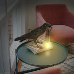 A black crow-shaped lamp stands on two books, holding a light-emitting device in its mouth, and the light softly illuminates the books.