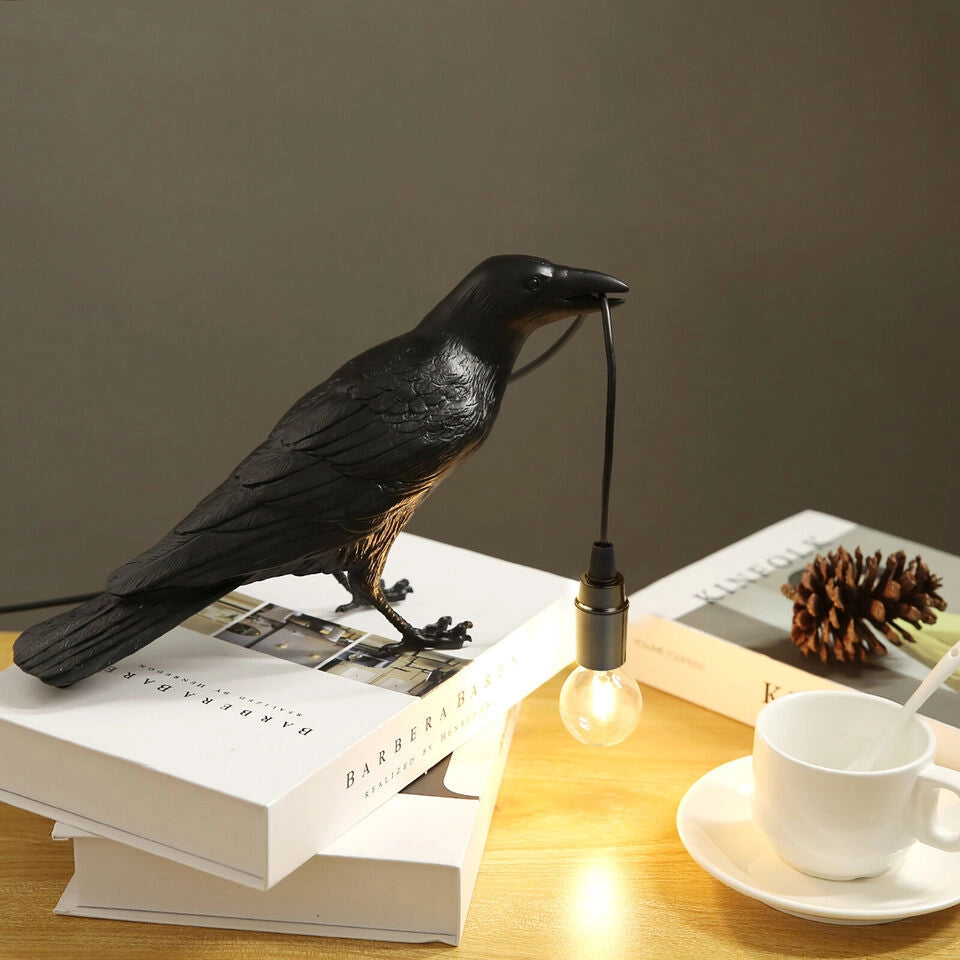 A black crow-shaped lamp stands on a stack of white books. A cup of coffee with a white spoon is placed on one side of the table, and there is a book next to it, with a pine cone on the book, creating a warm and quiet atmosphere as a whole.