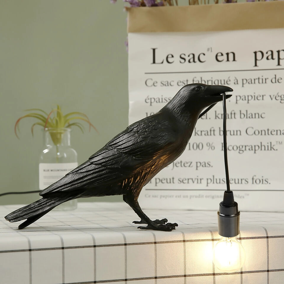 A black crow-shaped lamp stands on a checkered tablecloth, and the crow holds a black wire connected to the light bulb in its mouth