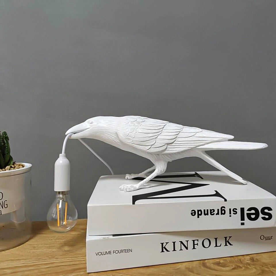 A white crow-shaped lamp, the light bulb emits warm light. The lamp is placed on a wooden table, and there is a pot of cactus in the background.