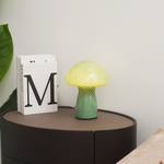Green and yellow table lamp on a round wooden table with books and a plant.