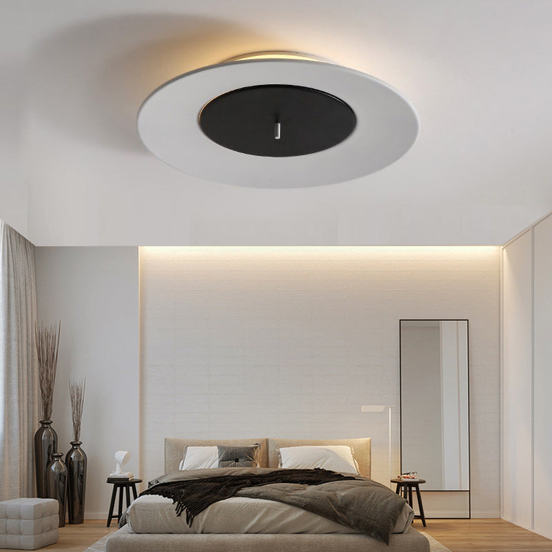 In a minimalist bedroom, a white round LED ceiling lamp hangs from the ceiling, with the black area in the middle contrasting with the soft warm light at the edge.