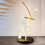 A graceful white female sculpture intertwined with golden curved LED arcs forms this artistic floor lamp, featuring spherical light accents on a sleek black base.
