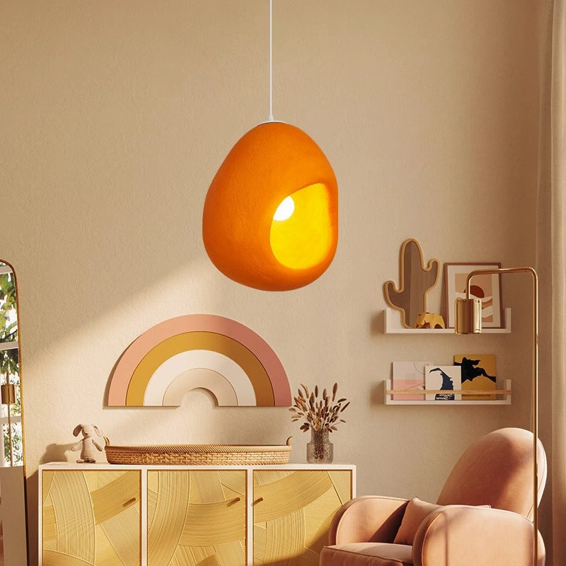single warm orange sensi pendant light hanging above rattan sideboard, boho kids room with rainbow wall decor and fabric armchair