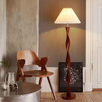 The picture shows a corner of the room. The spiral wooden column floor lamp emits warm light and is matched with a white pleated lampshade. Next to it are wooden chairs, a metal round table (with decorations on it), and a black-framed art painting. The wall is a combination of cement gray and dark gray. The overall style is simple and artistic.