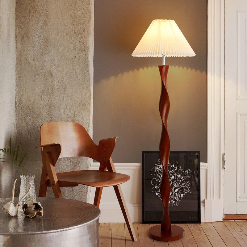 The picture shows a corner of the room. The spiral wooden column floor lamp emits warm light and is matched with a white pleated lampshade. Next to it are wooden chairs, a metal round table (with decorations on it), and a black-framed art painting. The wall is a combination of cement gray and dark gray. The overall style is simple and artistic.