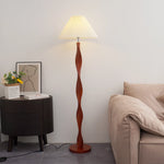 The dark brown walnut lamp post is in a twisted spiral shape. The solid wood texture rises and falls naturally with the arc. Each circle of spiral pattern is like a hand-carved annual ring. The interweaving of light and shadow reveals a warm texture. The white pleated lampshade on the top is like a blooming paper flower. The pleated texture is delicate and soft. When the warm light passes through, it casts a wavy halo on the wall. The metal base at the bottom of the lamp post