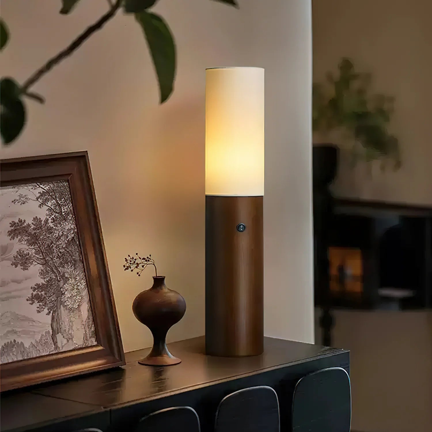 Lit Wooden Pillar Floor Lamp with dark wood base and fabric shade sits on dark cabinet beside framed art